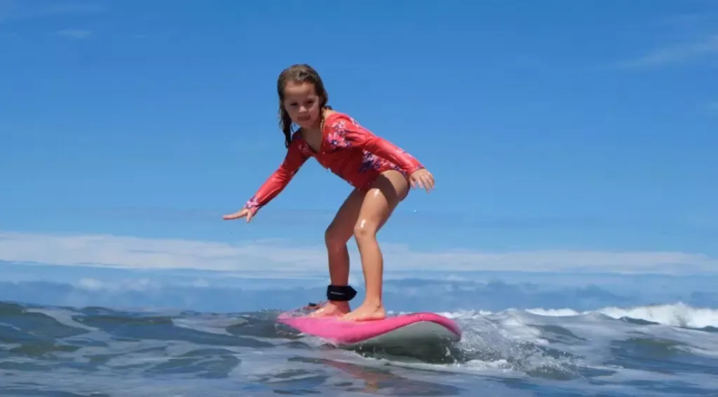 Joven surfer sobre una tabla rosa, de rodillas, remando en olas azules bajo cielo claro.