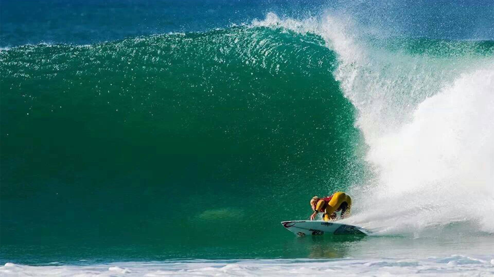 Surfer crouches low on a shortboard riding a massive green barrel, with white water spray behind in a breaking ocean wave.